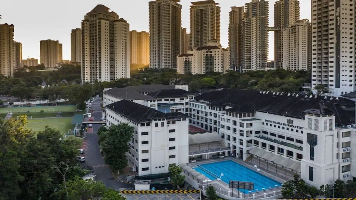 Aerial view of Mont'Kiara International School campus with swimming pool, sports field, and Kuala Lumpur skyline behind.