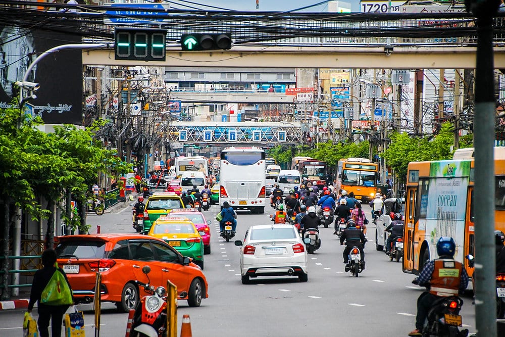 Heavy traffic on a busy Bangkok street with taxis, buses, motorcycles and overhead pedestrian bridges.